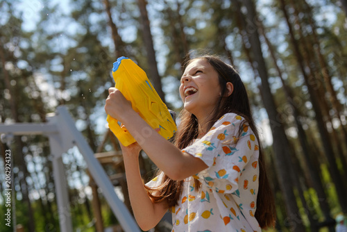 A girl holds a yellow water gun and laughs while playing outside. She is surrounded by tall trees in a sunny area during summer
