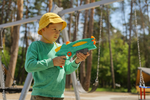 A child stands in a park holding a colorful water toy. The boy wears a yellow hat and green shirt while playing near swings on a sunny day