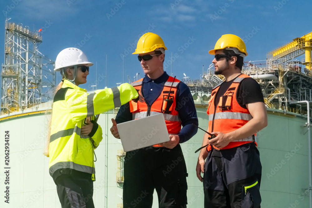 Fototapeta premium Workers discuss project at industrial site during daytime with machinery and storage tanks in background