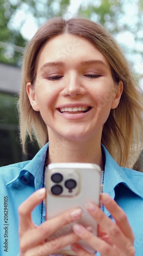 Close-up of satisfied young woman face with long hair and white teeth looking at mobile phone with pleased smile, standing over modern building background