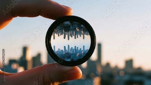 Round optical lens held in fingers inverting urban buildings.