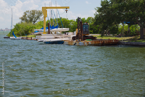 Fotografie Sailboats with blue covers lined up on trailers beside a marina dock, waterfront water in Gulfport, FL the foreground and trees in the background conveying leisure, calm and summer boating lifestyle