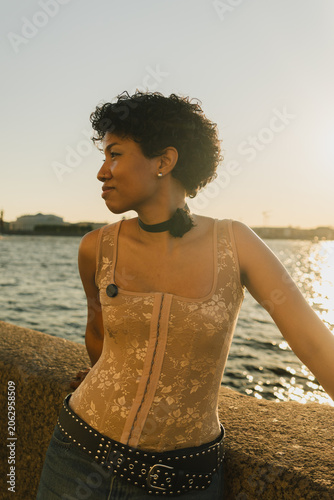 African American woman with curly hair stands by water, looking away. Wears beige corset top, black choker, studded belt. Golden hour light. Serene, fashion, outdoor. High quality photo