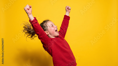 Energetic Girl in Red Sweatshirt Cheering with Arms Raised on Yellow Background