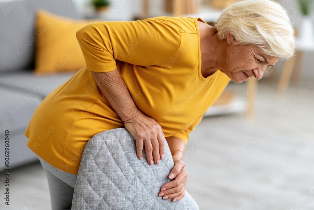 Fototapeta premium Elderly woman experiencing hip pain while trying to stand up from a chair, showing discomfort and agony.