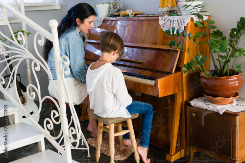 A young student receives a piano lesson from a female instructor in a cozy home environment. The room features plants and an antique piano.
