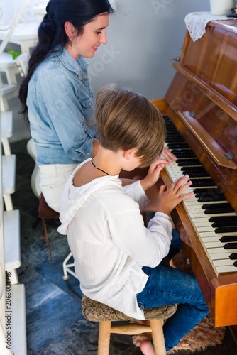A woman gently guides a child as they learn to play the piano. The child sits on a small stool, concentrating on the keys while receiving instruction.