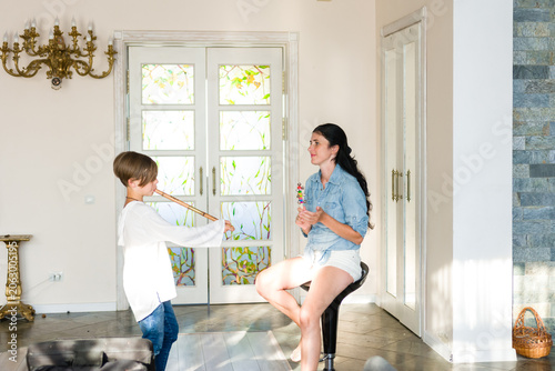 A girl plays a flute for her mother, who sits and listens attentively. The setting is a spacious, sunlit room with large windows and elegant decor. A heartwarming scene of family connection.