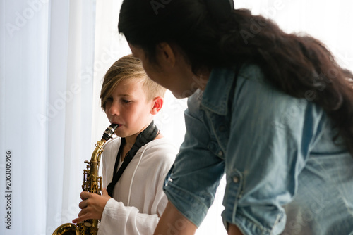 A boy, possibly 8-10 years old, practices the saxophone. An adult woman looks on, offering guidance or support. The setting appears to be a home, near a window.