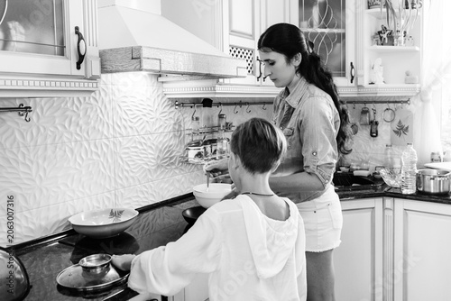 A grayscale image captures a heartwarming scene of a woman guiding a child in cooking. The kitchen is bright and modern, suggesting a comfortable and happy home environment.