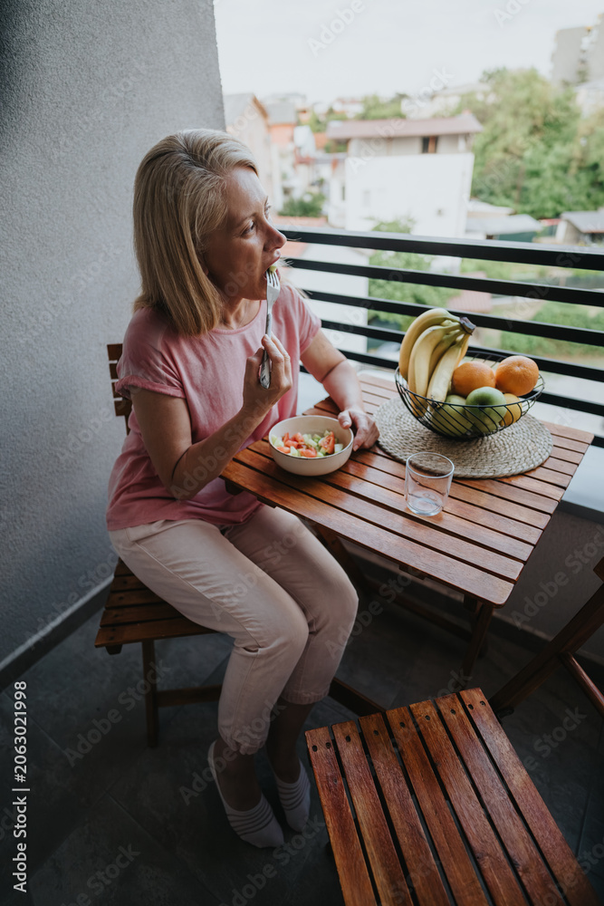 Fototapeta premium A middle-aged woman sits on a small balcony eating a bowl of salad. She has a light meal at a wooden table beside a bowl of fruit overlooking the neighborhood.