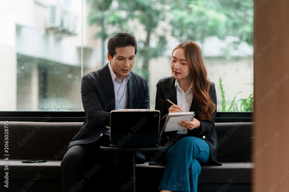 Obraz premium Two Asian business colleagues reviewing data on a tablet and taking notes, discussing strategy and working together in an office setting