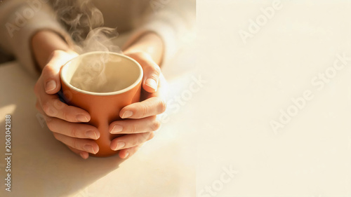Close up of two hands holding a steaming brown ceramic mug filled with hot coffee or tea on a light surface in warm lighting.