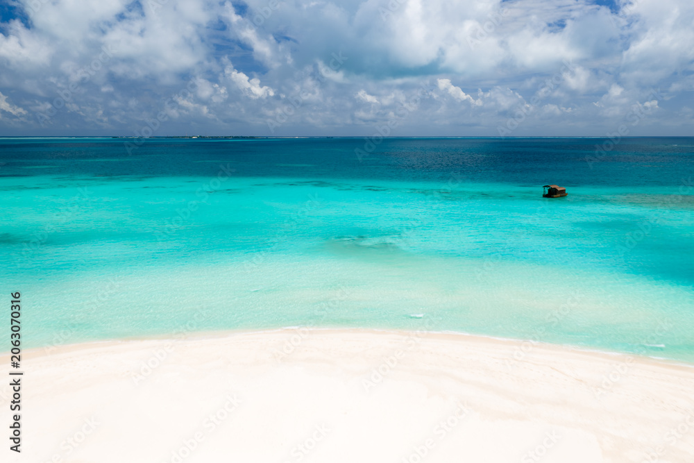Fototapeta premium White sand arcs into shallow turquoise water deepening to sapphire. A small buoy sits offshore. Low islands line the horizon under soft daylight and cumulus clouds.