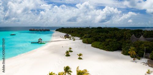 Obraz na plátně Aerial view of a Maldives resort with a curving white sand beach, a wooden jetty to an overwater pavilion, water villas above a lagoon, palm trees, and bright daytime clouds