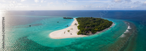 Obraz na plátně Aerial view of a Maldives island resort with a long sandbar, overwater villas on a jetty, palms, thatched umbrellas, anchored boats, and gentle surf on a sunny afternoon