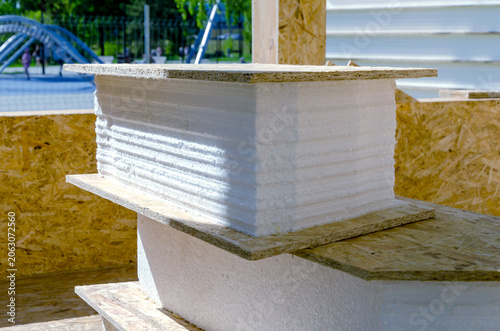 Stacked white foam insulation panels and OSB boards at construction site. These materials are used for SIP panel frame house construction. Proper storage ensures quality building materials.