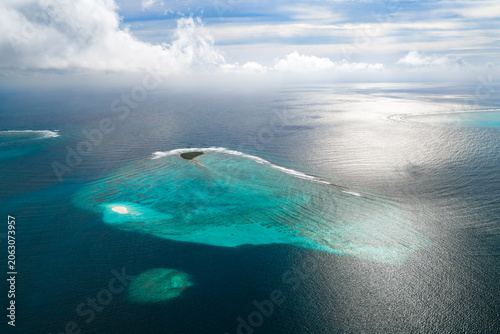 Foto Aerial view shows a small uninhabited sandbank and teardrop reef in the Maldives, with surf on the rim, bright aqua coral heads, soft high clouds, and midday light
