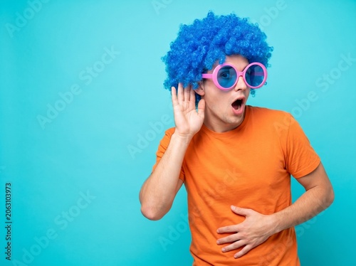 Humorous man sporting a vibrant blue afro wig and oversized pink eyewear actively listens intently, cupping his ear with one hand while expressing surprise and amusement with an open mouth, his