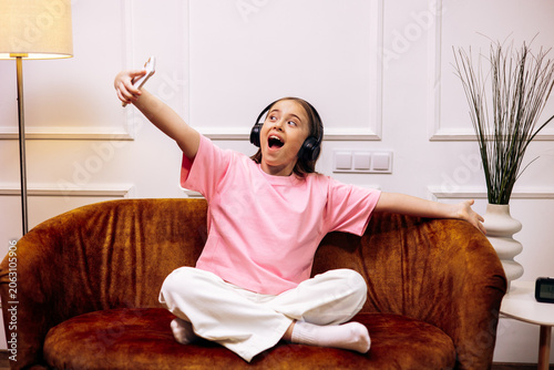 Young girl wearing headphones and pink t-shirt taking a selfie, singing along to music while relaxing on a couch at home