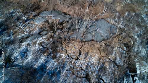 A drone shot looking down at a massive woody beaver dam blocking a ravine creek, creating a large frozen wetland pond surrounded by bare winter sunlit trees.