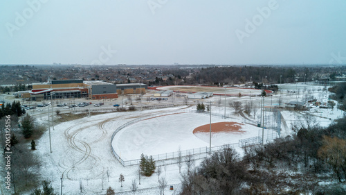 Fotografia A crisp high-angle shot capturing a snow-covered baseball field with its brown infield exposed, overlooking a high school campus, running track, and suburban park on an overcast winter day