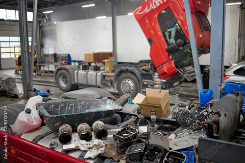 A detailed close-up of a disassembled engine in a machinery repair shop, with a raised cabin truck in the background. Tools and parts scattered around, showcasing the complexity of mechanical repairs.