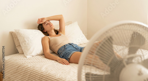 Overheated young woman lying on bed suffering from summer heatwave using an electric fan to cool down at home