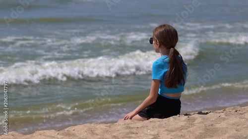 Young girl with long braided hair sitting alone on sandy beach surrounded by grass looking out over calm sea under cloudy sky