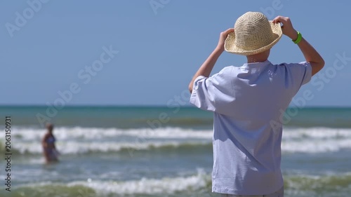 A teenager wearing a hat stands on a tropical island beach, admiring the sunset. A peaceful summer getaway or vacation; summer travel concept.
