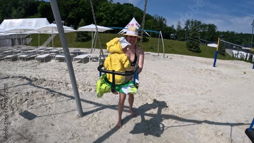 Woman pushing child on swing at sandy beach playground