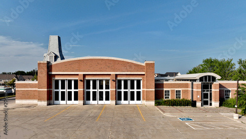 Fotografie Aerial view of a generic brick fire station featuring large bay doors and arched entrance in a residential neighbourhood backdrop on a sunny summer day with a clear blue sky