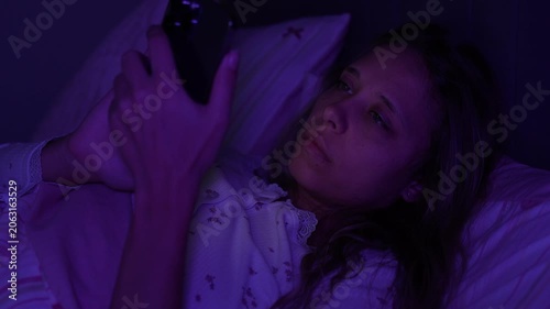 Young woman lies awake in bed at night using smartphone in purple light. Insomnia, social media addiction, blue light and sleep problems concept