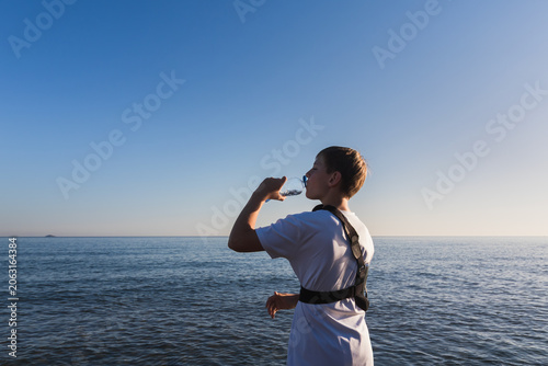 Boy with a broken arm wearing a medical shoulder sling brace drinks water from a bottle on the beach, focusing on hydration during summer recovery