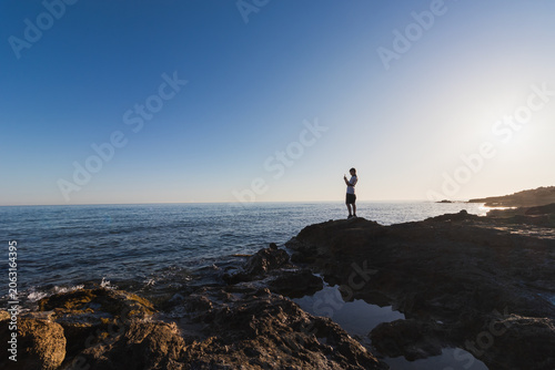 Silhouette of an 11 year old boy standing on a rocky coast in Crete and taking a picture of the sea with his smartphone