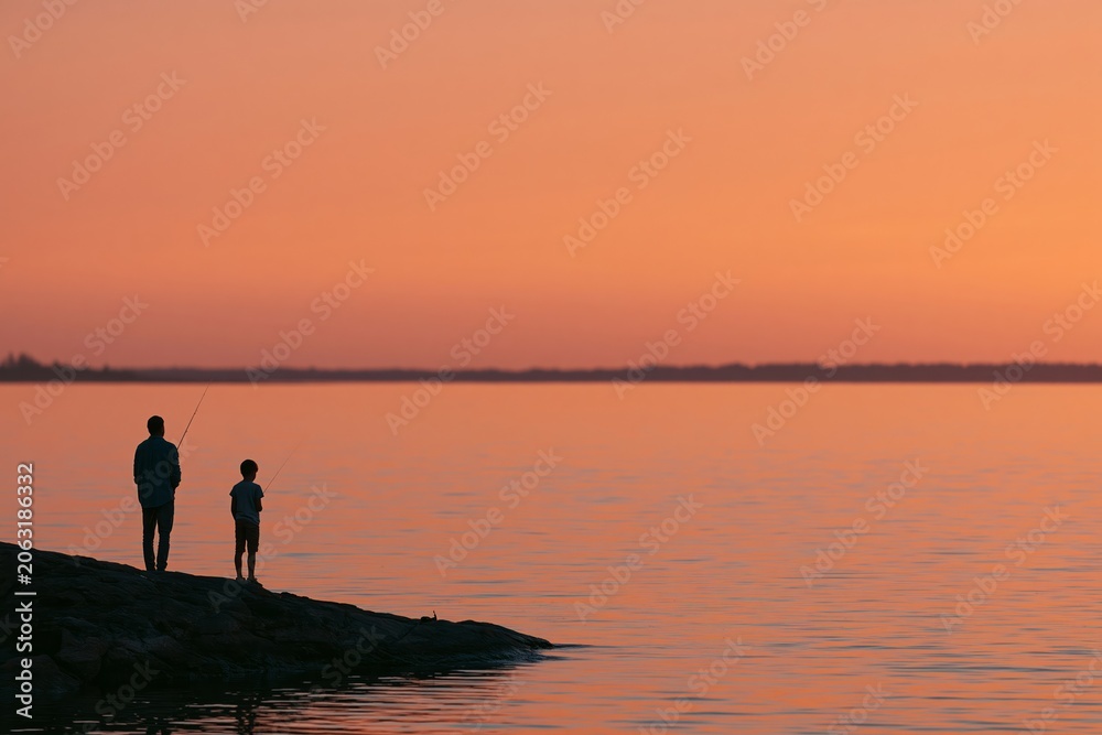 Fototapeta premium Father and son silhouette fishing together at sunset on a rocky shoreline, calm water reflecting warm colors of the evening sky