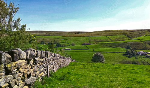 A picturesque landscape featuring a dry stone wall that stretches across the verdant countryside. The scene captures the undulating, green hills under a clear blue sky near, Haworth, UK