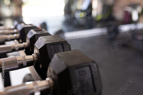Hexagonal rubber coated dumbbells are resting on metal rack in gym, chrome handles reflecting light