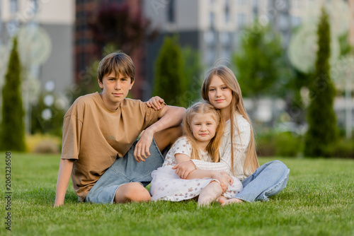 three children sitting on a lawn in the summer, in front of a residential complex in a city park