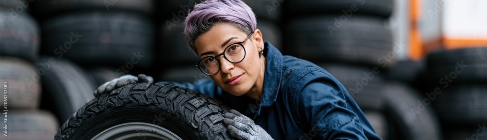 Obraz premium Portrait of a mechanic with purple hair leaning over a tire in a workshop background with stacked tires for industrial repair poster and banner design