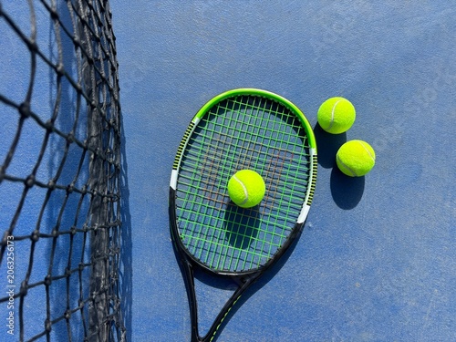 Horizontal flat lay of a tennis racket on a blue court with three tennis balls arranged around it, one resting on the strings. The tennis net is visible in the background.