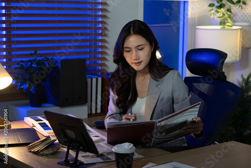 A focused Asian business professional examines a design catalogue at her desk during a late-night shift.