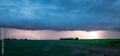 Dramatic lightning storm illuminates a rural field and lonely farmhouse beneath low, menacing thunderclouds at dusk.