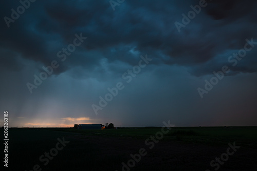 Dramatic dark storm clouds gather over a plain rural farm landscape at dusk, with distant light on the horizon