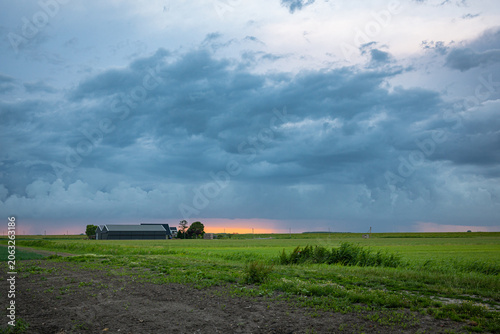Dark storm clouds sweep across a rural farm field at sunset, a dramatic blue-gray sky above a green field and distant barn.