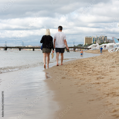 A young Caucasian man and a young woman with dark hair walk barefoot along a sandy beach