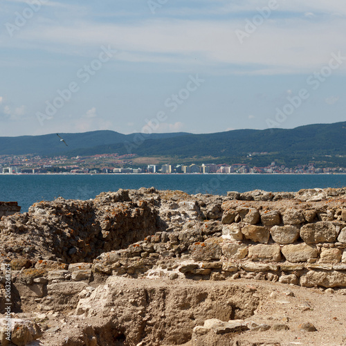 Ancient ruins by the sea with a distant city skyline and mountains in background.