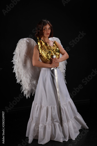 Full length cinematic portrait of a beautiful woman in a golden corset and white angel wings holding a medieval sword on a black background