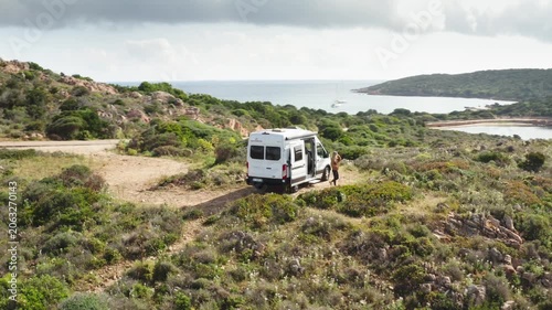 Wide Drone shot of girl walking out of Camper van on mediterranean island as drone circles van overhead
