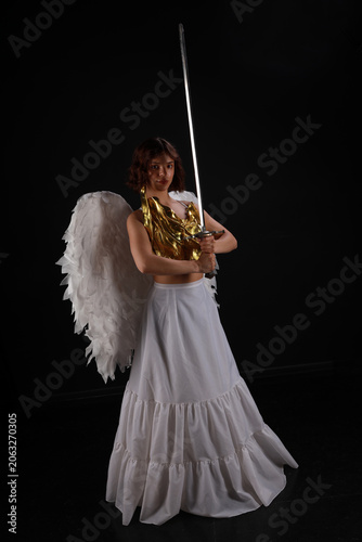 Full length cinematic portrait of a beautiful woman in a golden corset and white angel wings holding a medieval sword on a black background